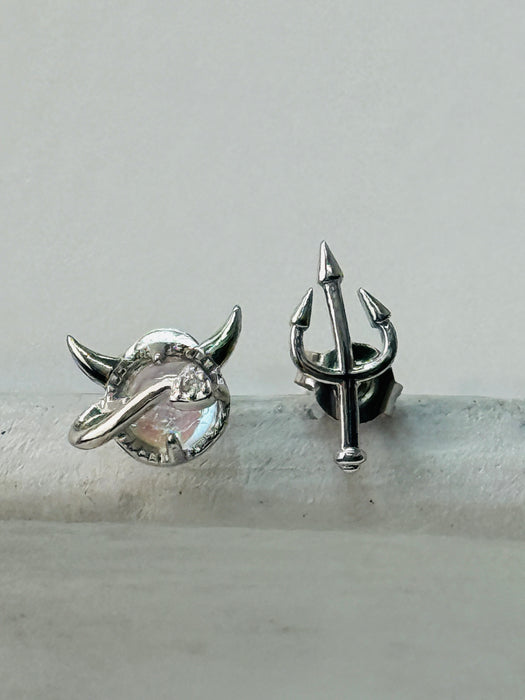 A pair of silver-toned stud earrings with one featuring a devil horned planet design and the other with a trident design.