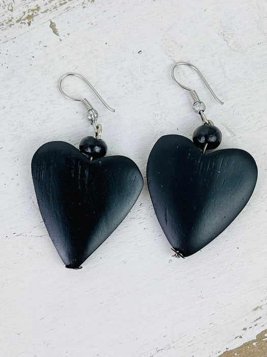 A pair of black heart-shaped wooden bead earrings with hook closures on a white wooden surface.