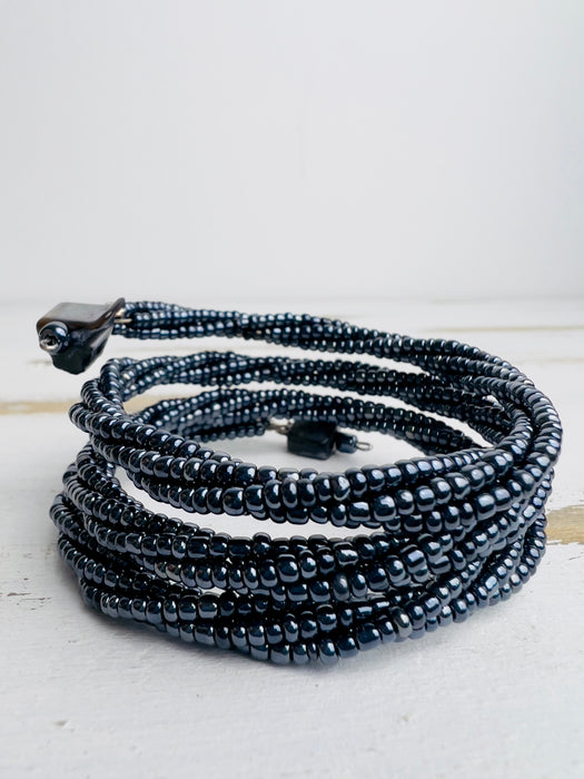 A gunmetal-coloured seed bead bracelet with a plaited effect and shell detail at each end, displayed on a wooden surface.