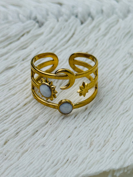 Gold celestial harmony ring with crescent moon, star, sun and moonstone-effect cabochons on an open band, displayed on a white fabric background.