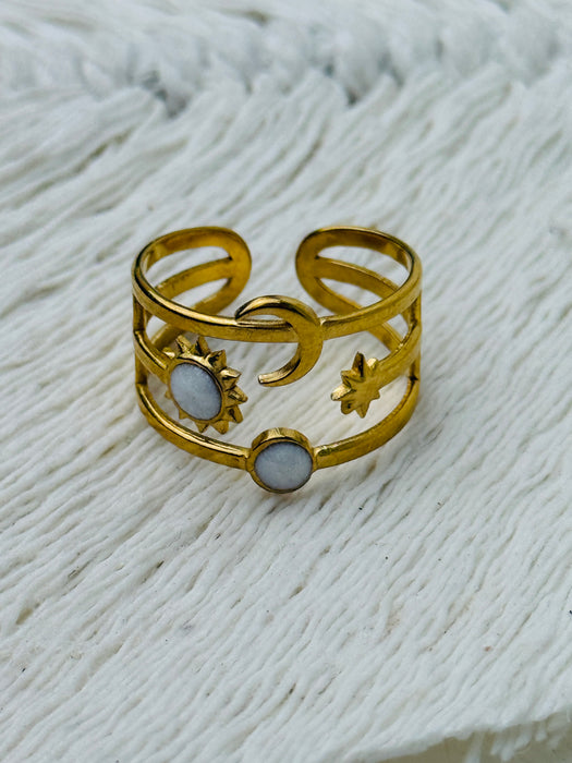 Gold celestial harmony ring with crescent moon, star, sun and moonstone-effect cabochons on an open band, displayed on a white fabric background.