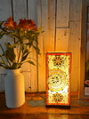 Handmade square mosaic lamp with a sunburst pattern, painted in whites and gold tones, placed on a wooden surface with a vase of orange flowers in the background.