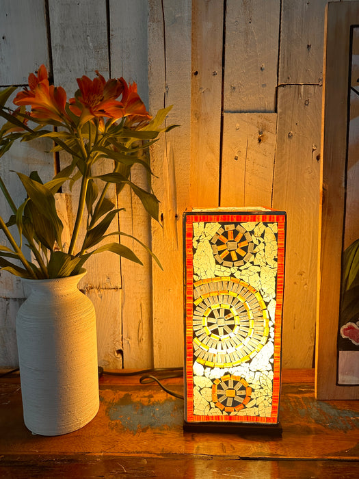 Handmade square mosaic lamp with a sunburst pattern, painted in whites and gold tones, placed on a wooden surface with a vase of orange flowers in the background.