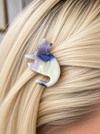 A small cat-shaped hair clip with a marbled purple, blue, and iridescent white finish, shown pinned to blonde hair.
