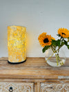 display view of mosaic cylinder lamp with light on placed on a wooden surface next to a vase of sunflowers