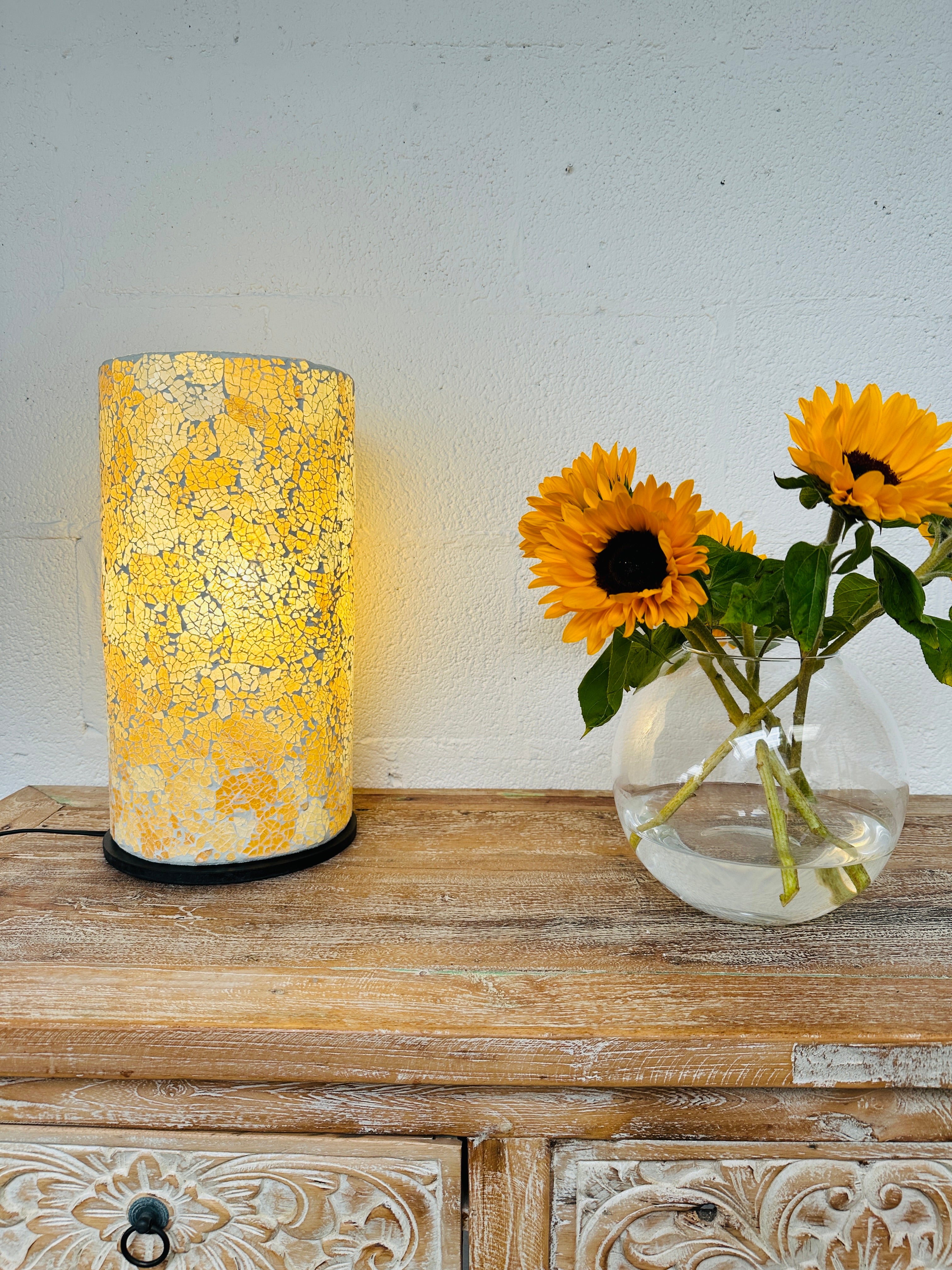 display view of mosaic cylinder lamp with light on placed on a wooden surface next to a vase of sunflowers