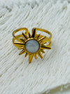 Gold sunburst ring with moonstone-effect acrylic cabochon on open band, displayed on a fabric background.
