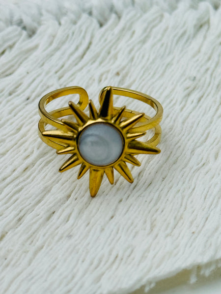 Gold sunburst ring with moonstone-effect acrylic cabochon on open band, displayed on a fabric background.
