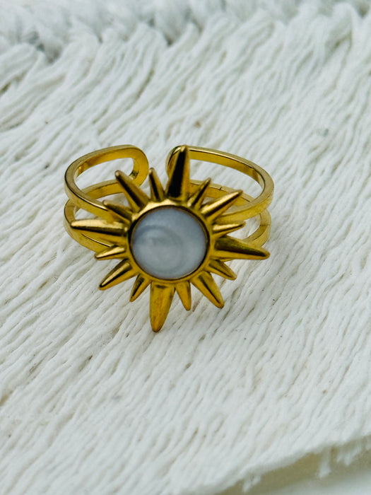 Gold sunburst ring with moonstone-effect acrylic cabochon on open band, displayed on a fabric background.
