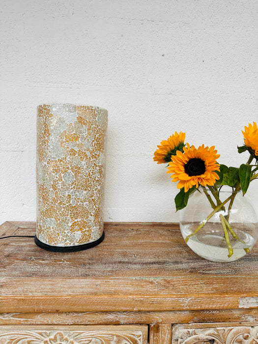 display view of mosaic cylinder lamp with light off placed on a wooden surface next to a vase of sunflowers
