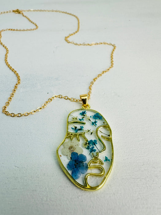 A gold-plated chain necklace with an abstract portrait pendant featuring pressed turquoise flowers set in resin.