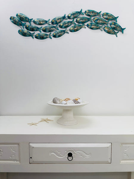 Mottled aqua metal fish shoal wall art displayed above a white side table with shells