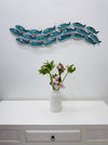 Aqua striped metal fish shoal wall sculpture displayed above a white cabinet with a vase of flowers