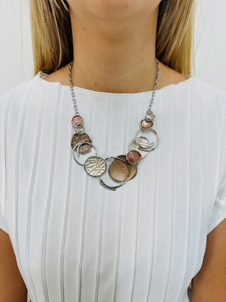 A beige enamel statement necklace with metallic accents and textured metal discs, displayed on a model.
