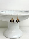 Pair of black and gold flower-shaped earrings on a white cake stand.