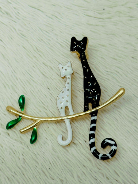A brooch featuring two stylized cats in black and white colors, with a gold-colored branch and green leaves.