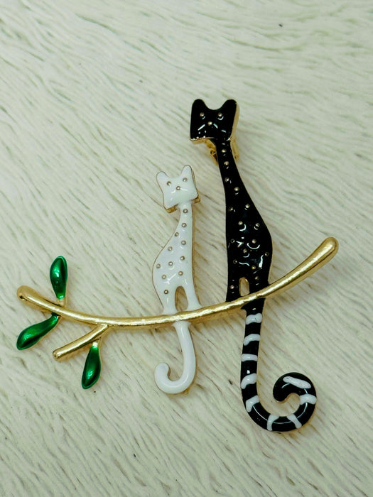 A brooch featuring two stylized cats in black and white colors, with a gold-colored branch and green leaves.