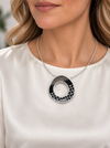A close-up of a woman wearing a modern silver-toned necklace with a textured circular pendant and a thin chain, paired with a cream satin top.