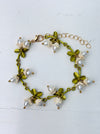 Gold-tone adjustable charm bracelet with white lily of the valley pearl effect drops and green enamel leaves displayed on a white background.