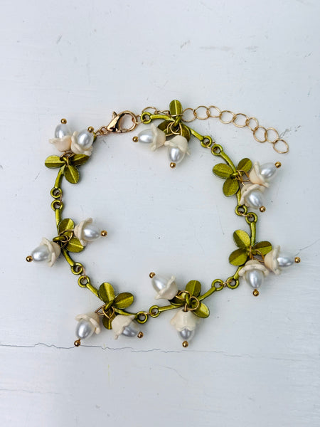 Gold-tone adjustable charm bracelet with white lily of the valley pearl effect drops and green enamel leaves displayed on a white background.