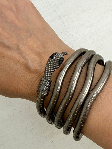 A close-up of a person's wrist wearing a stack of dark metallic snake-themed bracelets, including a prominent snake head clasp and coiled snake bangles.