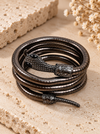 A coiled black metal bracelet with a detailed snake head clasp and textured scales, displayed on a beige stone surface with dried flowers.