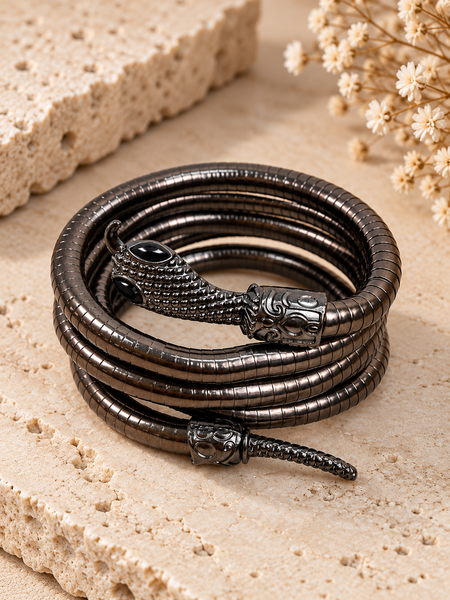 A coiled black metal bracelet with a detailed snake head clasp and textured scales, displayed on a beige stone surface with dried flowers.