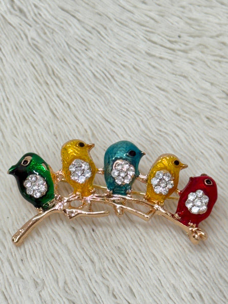 A brooch depicting a series of colorful birds sitting on a branch, with green, yellow, blue, and red colors, with the exception of the lead bird which is multi-colored with silver accents.