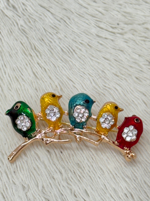 A brooch depicting a series of colorful birds sitting on a branch, with green, yellow, blue, and red colors, with the exception of the lead bird which is multi-colored with silver accents.