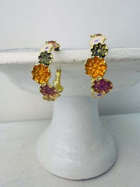 A pair of golden hoop earrings adorned with colorful enamel flowers displayed on a white stand.