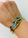An elegant gold cuff bracelet with deep blue and turquoise enamel leaf details, displayed on a person's wrist.