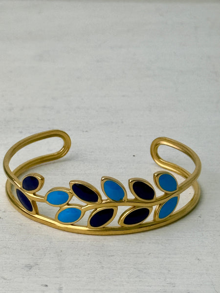 An elegant gold cuff bracelet with deep blue and turquoise enamel leaf details, displayed on a white background