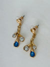 Gold-tone branch drop stud earrings with blue enamel teardrops and clear glass crystals on a white table.
