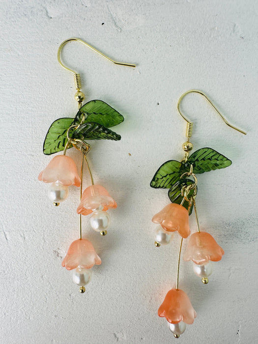 A pair of lilly dangle earrings with green leaves and orange flowers, with a pearl drop at the bottom.