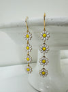 A pair of floral dangle earrings with white and yellow flowers on a gold-colored chain, displayed against a light background.
