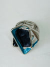 Close-up of silver-tone stretch ring with layered blue and gunmetal enamel geometric shapes.