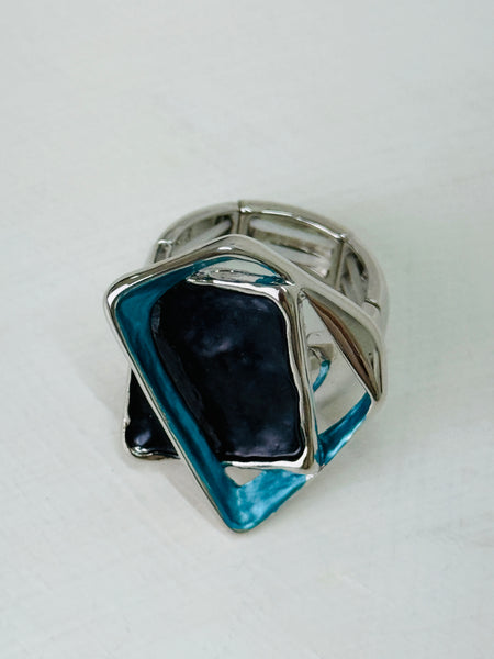 Close-up of silver-tone stretch ring with layered blue and gunmetal enamel geometric shapes.