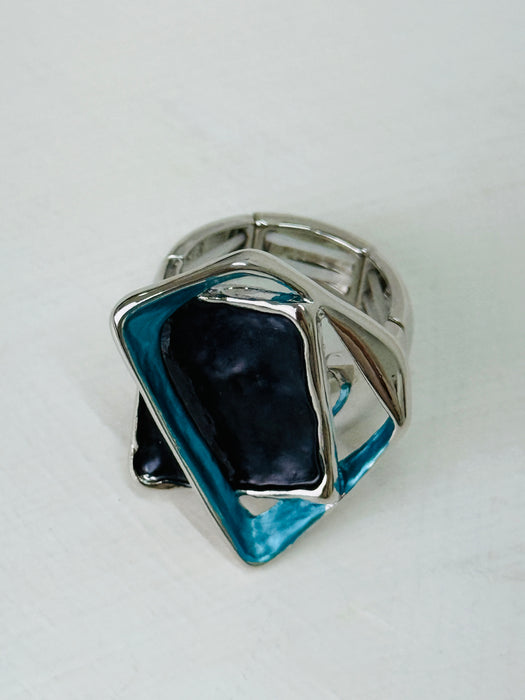 Close-up of silver-tone stretch ring with layered blue and gunmetal enamel geometric shapes.