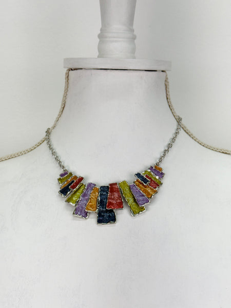 Multicoloured geometric statement necklace with rectangular enamel shapes in metallic gunmetal, bronze, gold, lilac and lemon green, displayed on a mannequin.