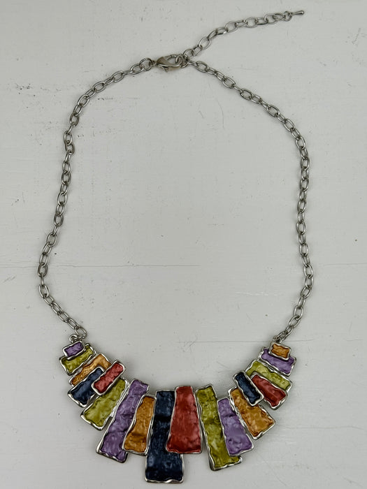 Multicoloured geometric statement necklace with rectangular enamel shapes in metallic gunmetal, bronze, gold, lilac and lemon green, displayed on a table.