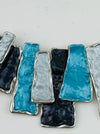 Close-up of a geometric necklace featuring rectangular enamel pieces in turquoise, grey, and gunmetal tones, with a silver-tone outline.