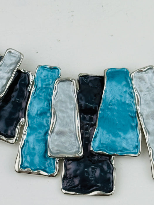 Close-up of a geometric necklace featuring rectangular enamel pieces in turquoise, grey, and gunmetal tones, with a silver-tone outline.
