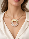 A close-up of a model wearing a modern circular pendant necklace. The necklace features a gold and silver-toned geometric ring with a textured, openwork lower half, suspended from a delicate gold chain.