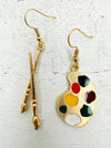 A pair of gold-colored earrings featuring a paint palette and paintbrush design.