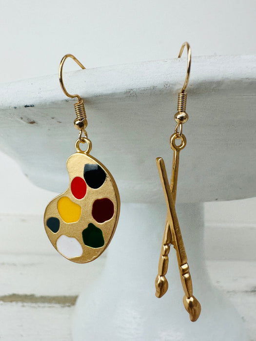 A pair of gold-colored earrings featuring a paint palette and paintbrush design.