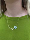 Necklace with a blue flower pendant worn by a person on a green shirt
