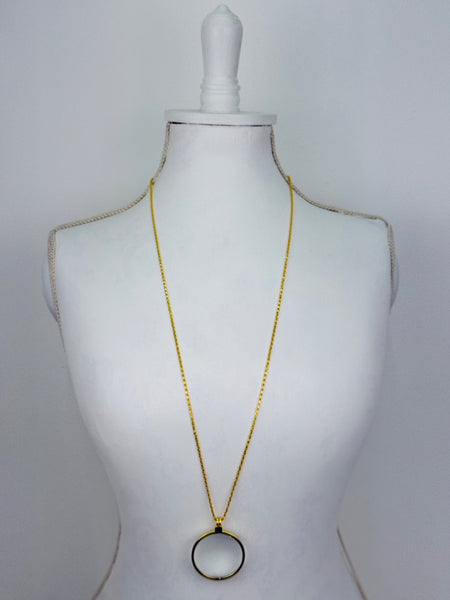 Gold magnifying glass pendant necklace with round glass lens on a gold chain.