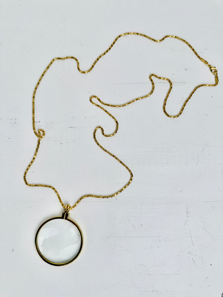 Gold magnifying glass pendant necklace with round glass lens on a gold chain, displayed on a white backgrond.