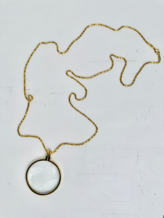 Gold magnifying glass pendant necklace with round glass lens on a gold chain, displayed on a white backgrond.