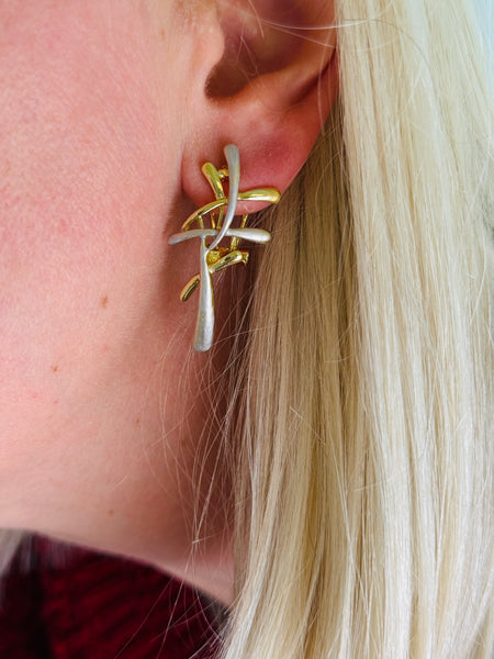 Close-up of a model wearing gold and silver tone abstract earrings with interwoven curved bars.